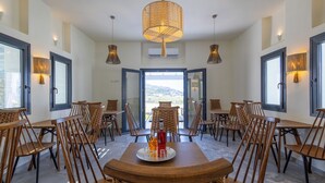 A dining area with wooden tables and chairs, a central table with a bottle and glass, and a view through the open door. - Cezaria Naxos Hotel (Naxos)