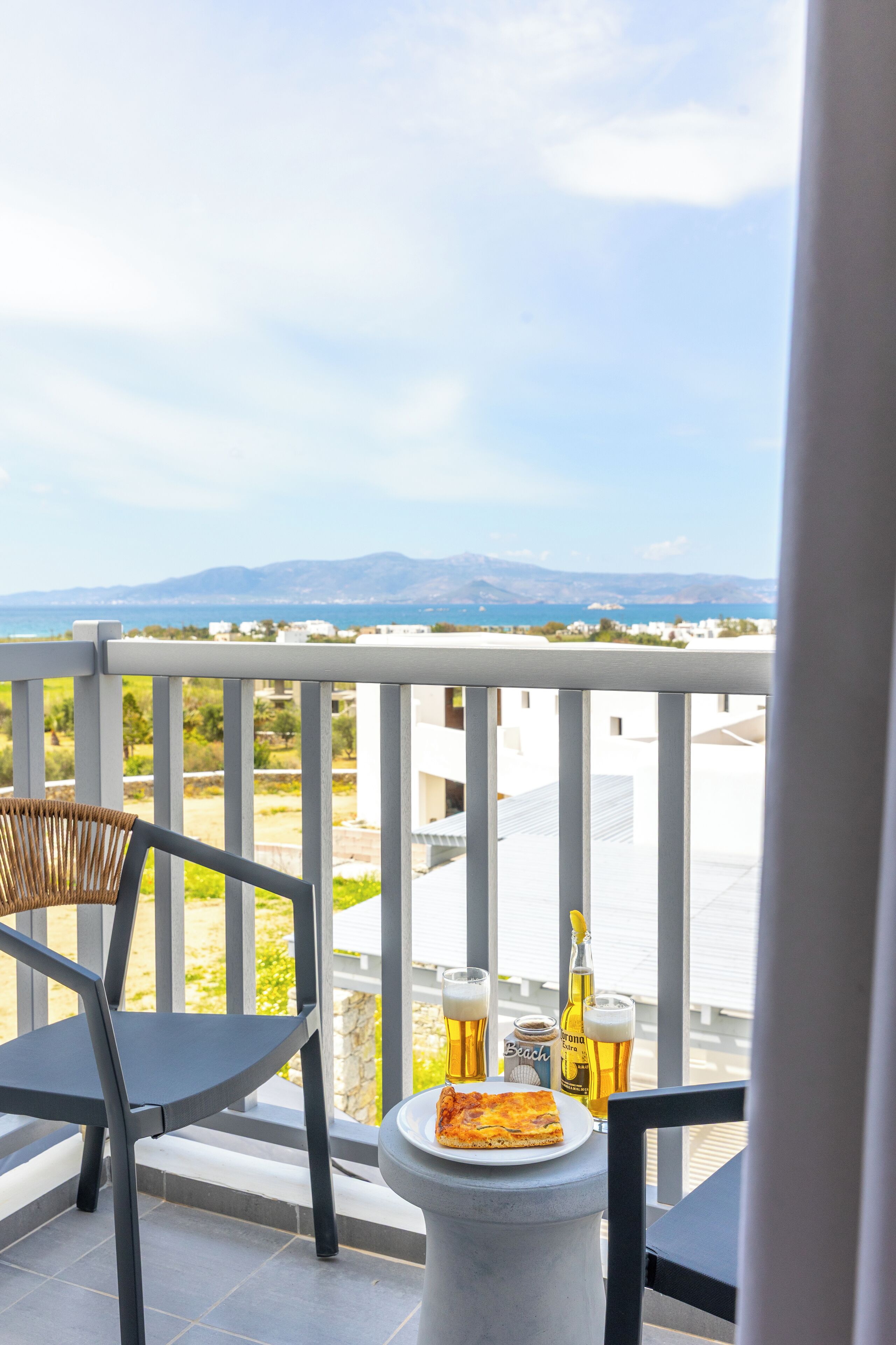 Ein Balkon mit Meerblick, ein Tisch mit einer Pizzascheibe und Biergläsern sowie ein Korbstuhl.