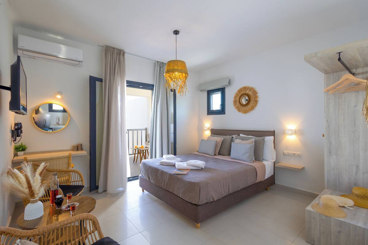 A modern bedroom with a large bed, a dining area with wicker furniture, and a balcony with a view.