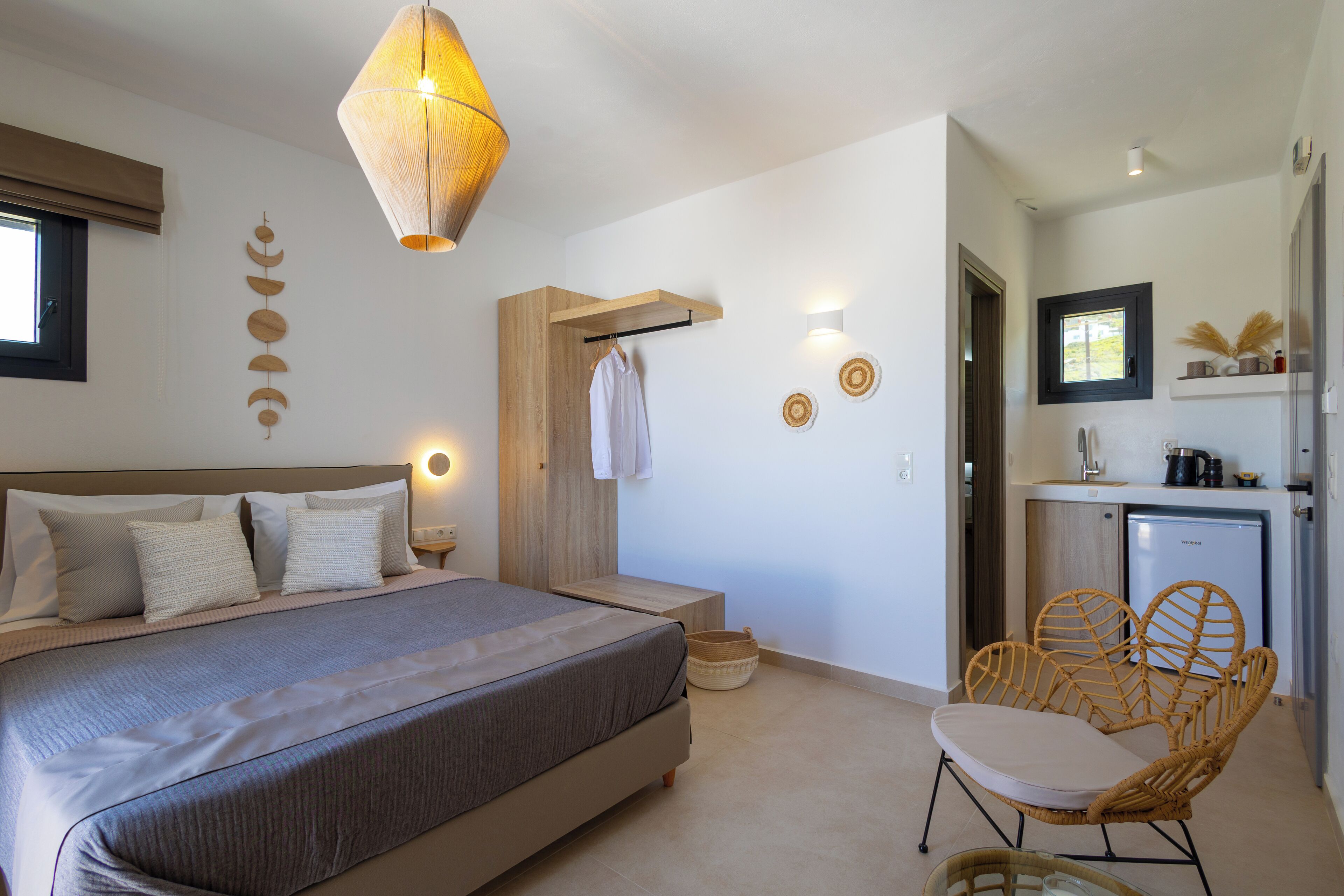 Un dormitorio con una cama grande, mesitas de noche, una lámpara colgante y una pequeña cocina americana con un refrigerador y una silla.