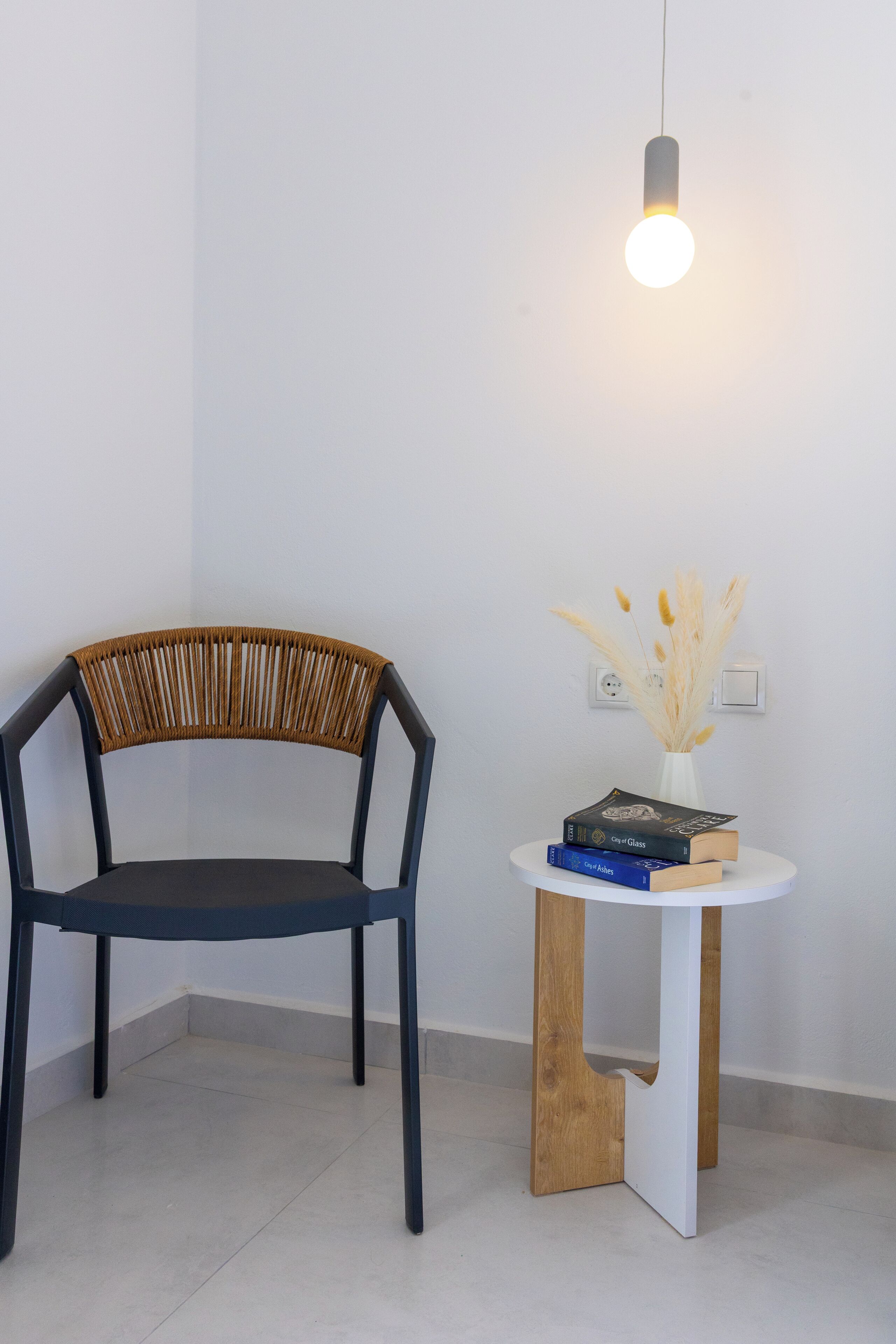 A modern chair with a wooden backrest and black seat, a small white table with a vase of dried pampas grass, and a hanging light fixture.