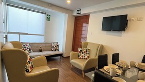 Habitación de hotel con sofá, sillones, una mesa de comedor con tazas y platos, un televisor montado en la pared y una ventana con persianas.