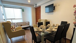 Un comedor con una mesa dispuesta para cuatro personas, rodeada de sillas, un sofá con cojines y un televisor montado en la pared.