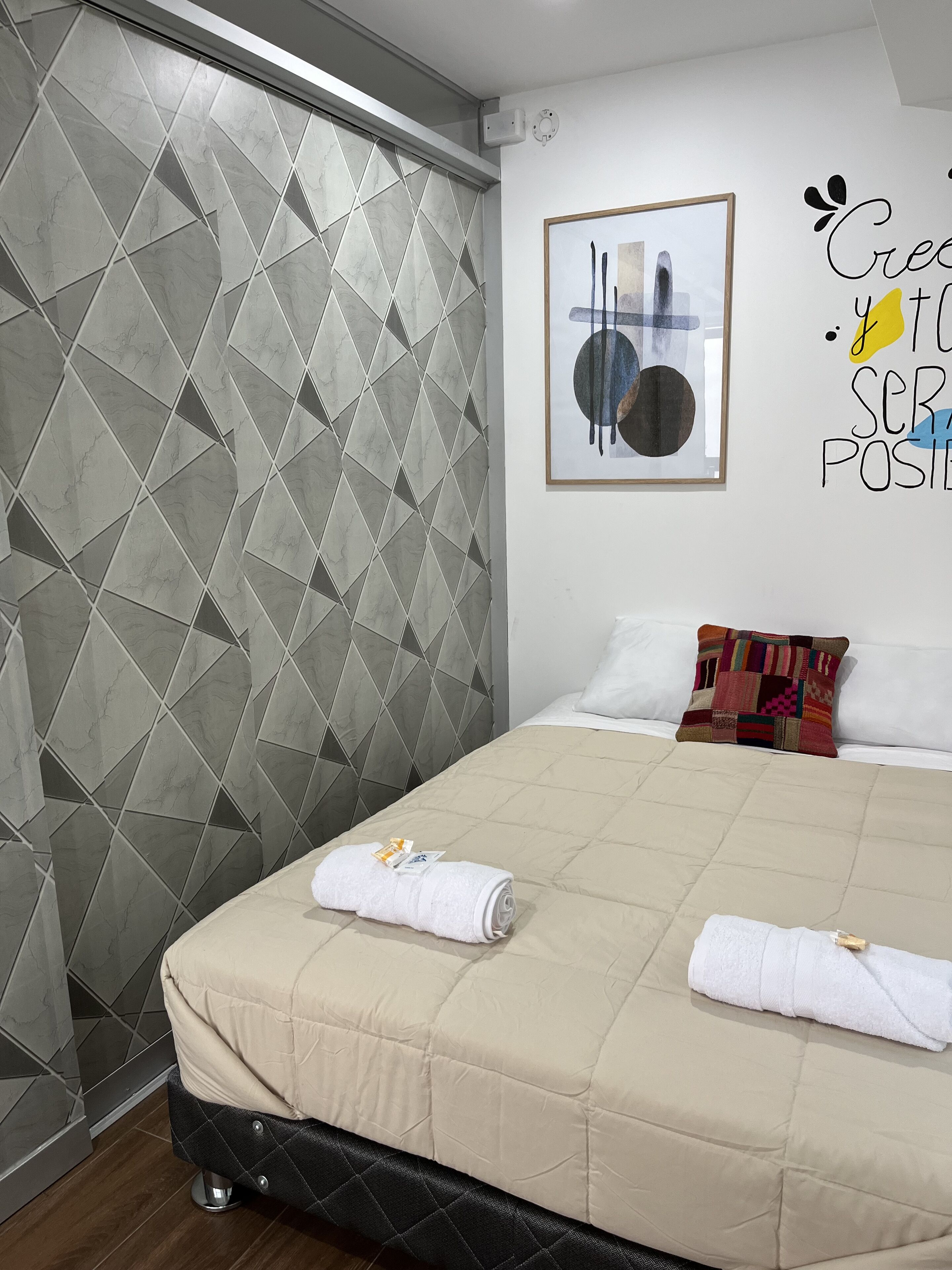 Une chambre d’hôtel avec un lit, une couvre-lit beige matelassé, une tête de lit noire recouverte de tissu, un coussin à motifs, une œuvre d’art encadrée au mur et un papier peint à motifs géométriques.