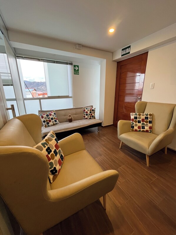 A room with two beige armchairs, a sofa, and a wooden floor. There is a window with a view of buildings and a door labeled "SALIDA".