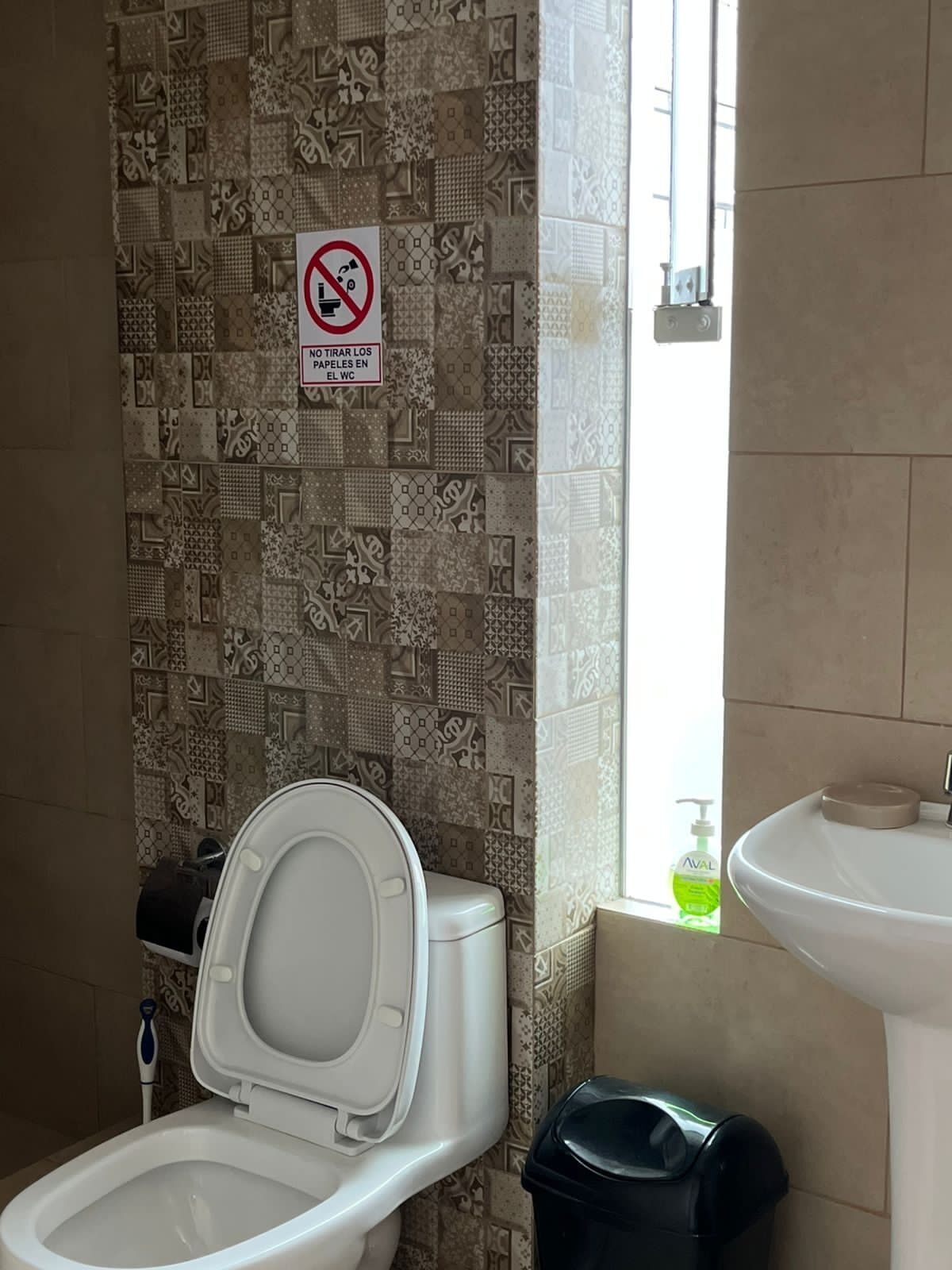 A bathroom with a patterned tile wall, a toilet, a trash bin, and a sink.