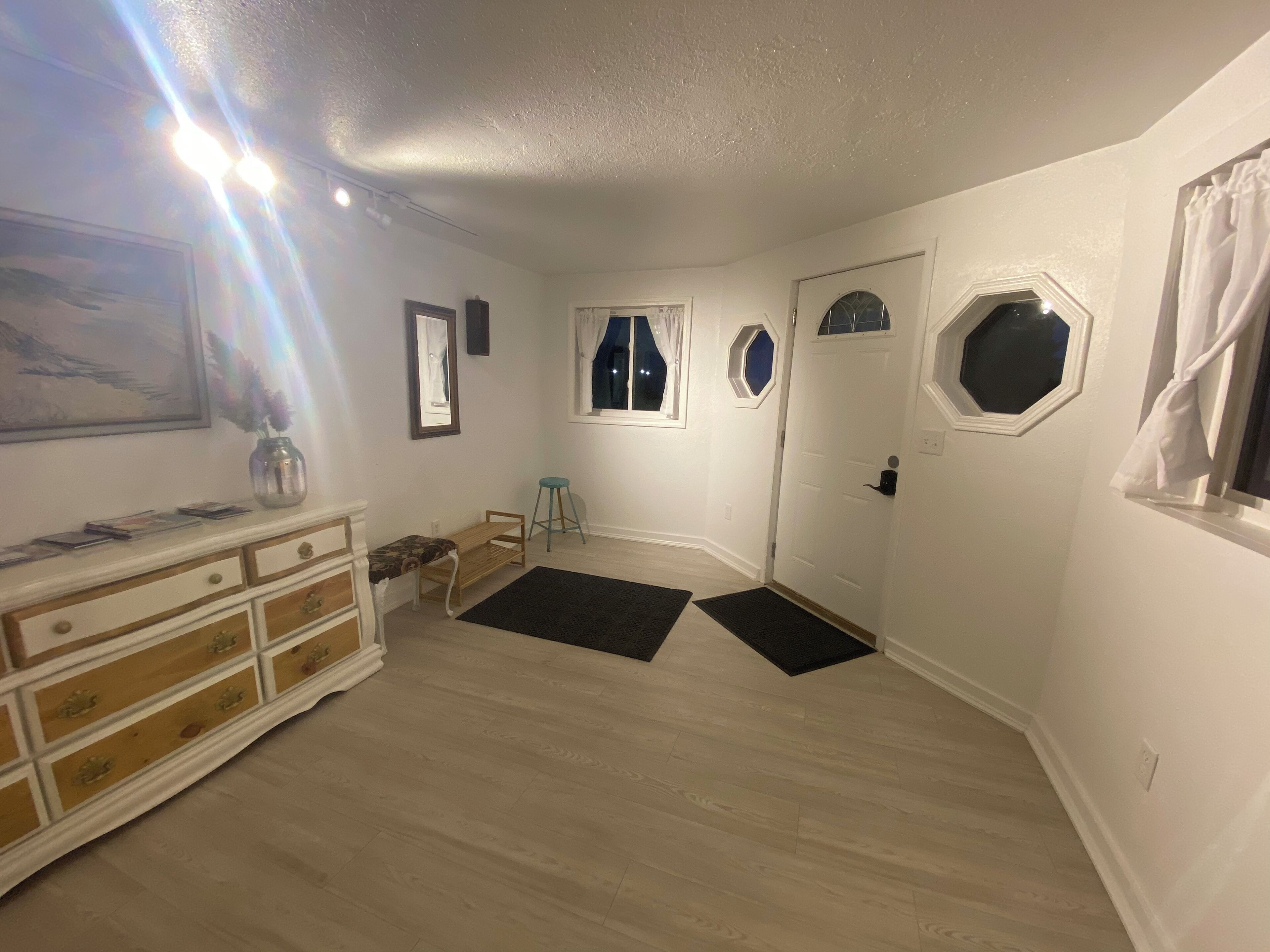 A room with a wooden dresser, a small stool, a door with a window, and a black rug on the floor.