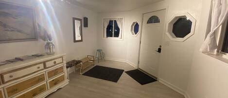 A room with a wooden dresser, a small stool, a door with a window, and a black rug on the floor.