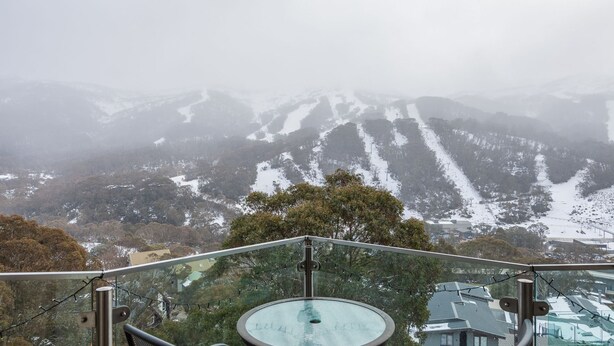 Une vue panoramique depuis un balcon doté d’une balustrade en métal, donnant sur un paysage montagneux aux pentes enneigées et traversé par une rivière.