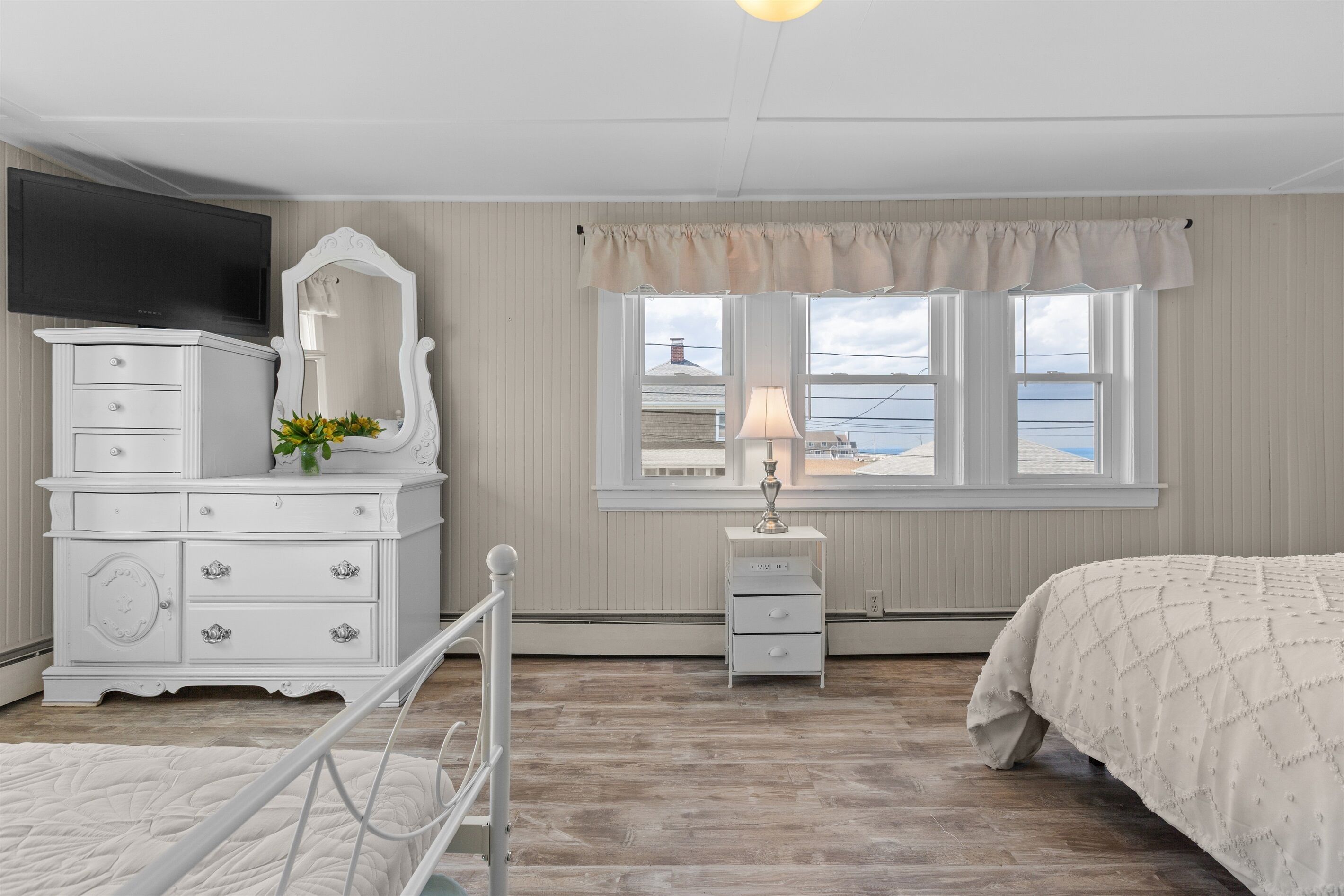 A bedroom with a bed, a dresser with drawers, a mirror, a bedside table with a lamp, and a window with a view of the sea.