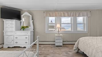 A bedroom with a bed, a dresser with drawers, a mirror, a bedside table with a lamp, and a window with a view of the sea.