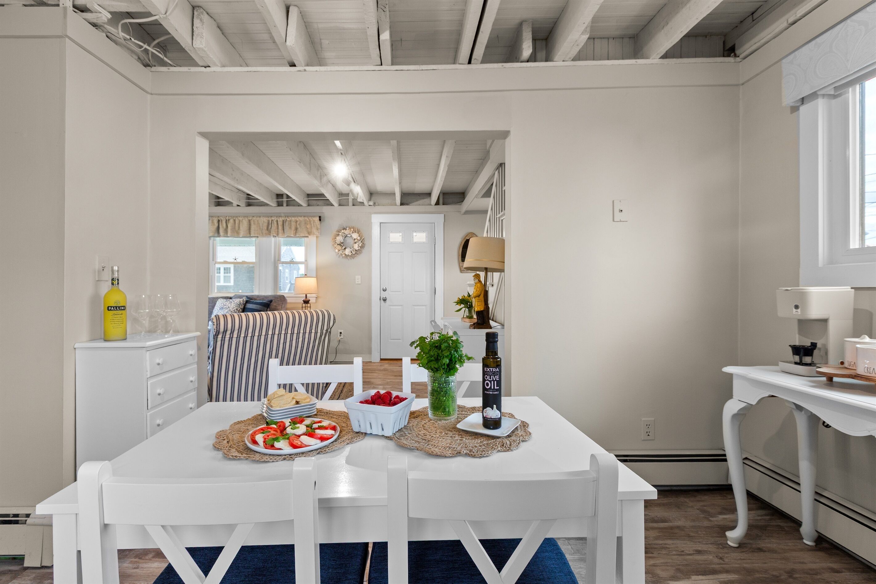A dining area with a white table set for two, a bottle of extra virgin olive oil, a basket of strawberries, and a small potted plant.