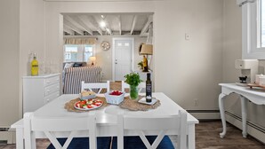 A dining area with a white table set for two, a bottle of extra virgin olive oil, a basket of strawberries, and a small potted plant. - Scituate Seaside Escape – Sleeps 8, Walk to Beach (Scituate)