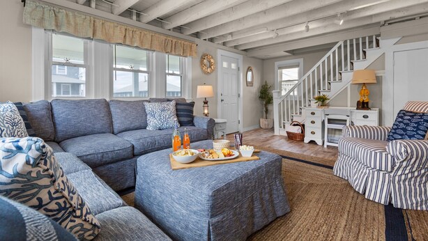 A spacious living room with a large sectional sofa, a striped armchair, and a wooden coffee table with snacks. A staircase with a white railing leads to an upper level. - Scituate Seaside Escape – Sleeps 8, Walk to Beach (Scituate)