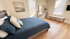 A bedroom with a bed, wooden floor, a dresser, a bench, and two windows with blinds. - Charming Central Home-3bd/1bath (Danville)