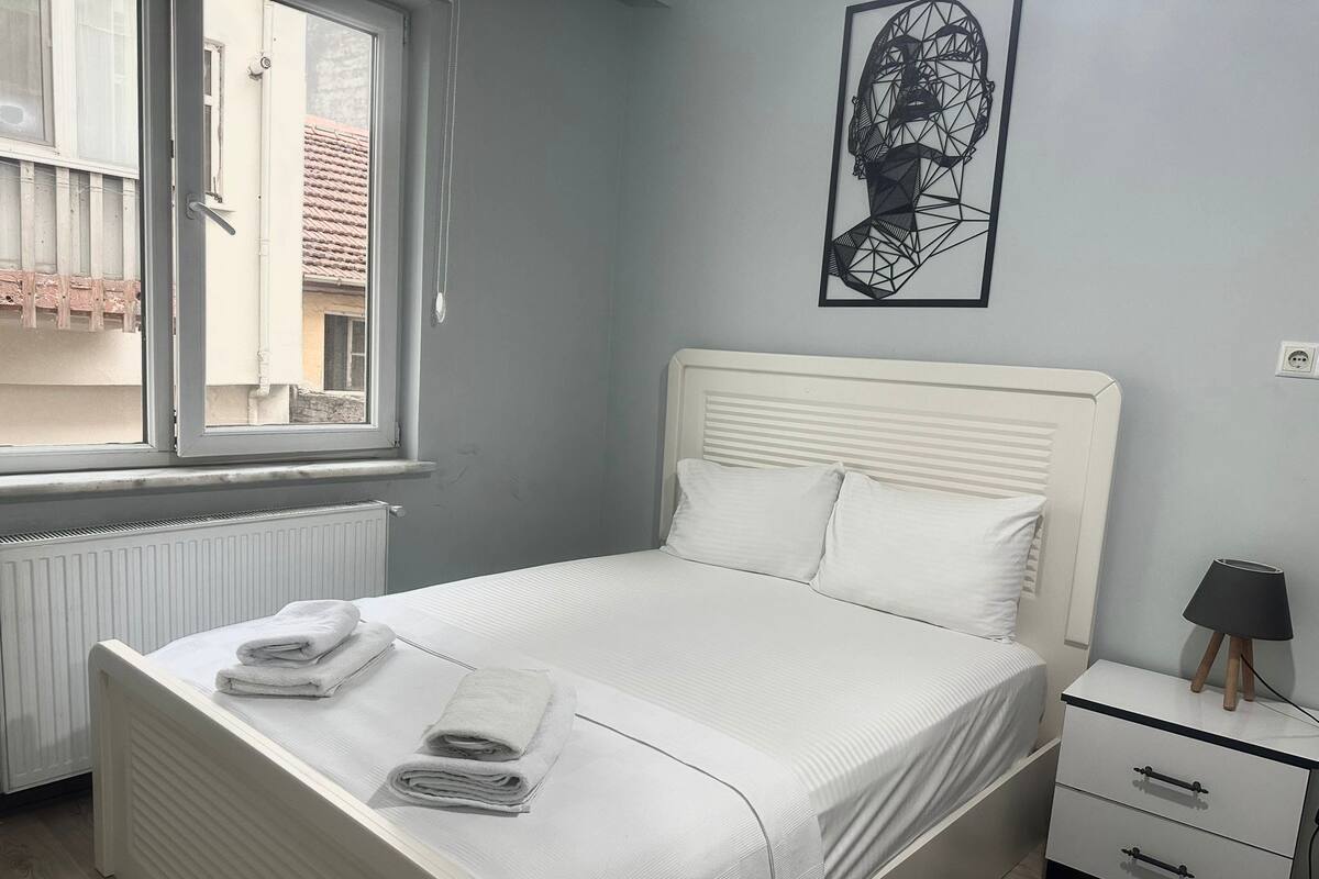 A bedroom with a bed, a nightstand, a lamp, a radiator, and a window.