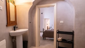 Une salle de bain avec un lavabo sur colonne, un miroir, une étagère contenant des articles de toilette et une porte donnant sur une chambre.