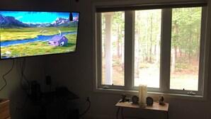 A bedroom with a TV on the wall, a window with a view of a forest, a bedside table with a lamp, and a bed with a red blanket.