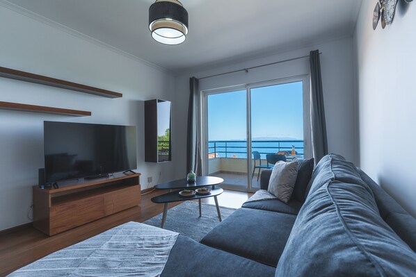 Un soggiorno moderno con una TV a schermo piatto, un divano, un tavolino da caffè e un balcone con vista sul mare.
