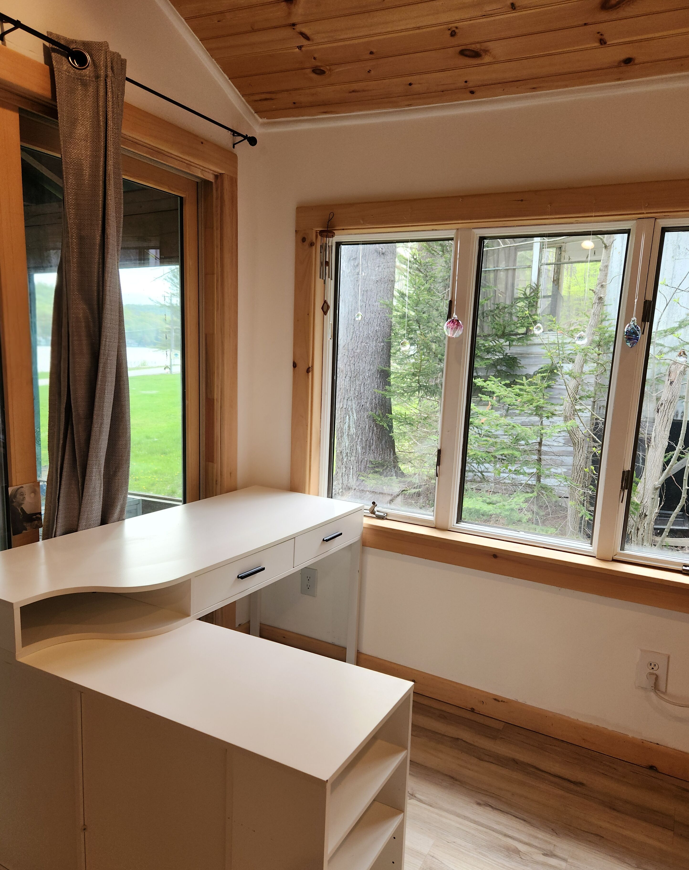 Una stanza con una scrivania bianca, una finestra vista alberi e un soffitto in legno.