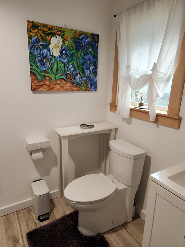 A bathroom with a toilet, a small trash bin, a white sink, and a window with curtains.