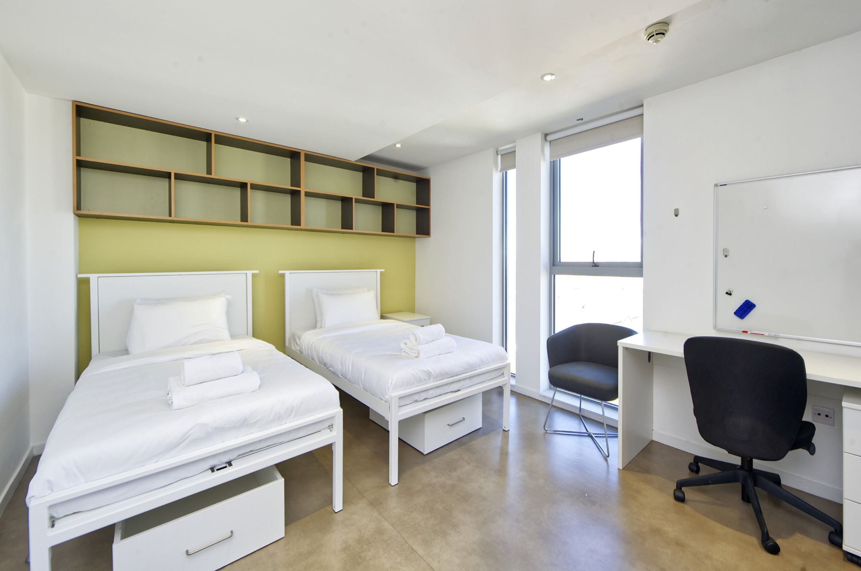 Habitación de hotel con dos camas individuales, un escritorio con silla, una pizarra y un ventanal grande.