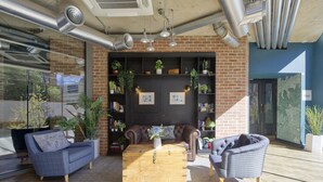 A modern lounge area with a leather sofa, a wooden coffee table, and a wicker chair. There are potted plants and framed pictures on shelves. - Comfort in Hammersmith Student Studio (London)