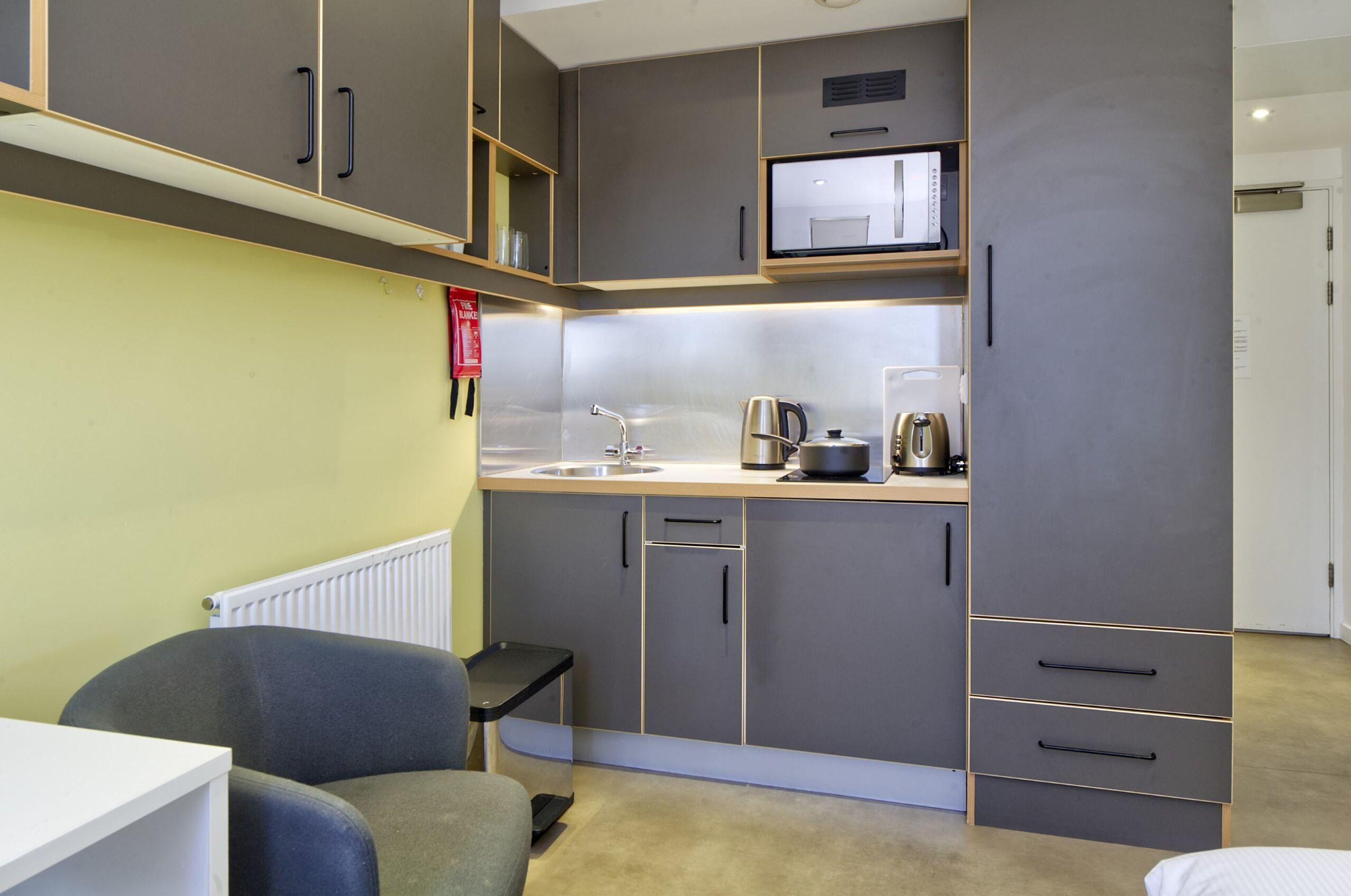 A compact kitchen with grey cabinets, a microwave, and a kettle. There is a small black chair and a white table.