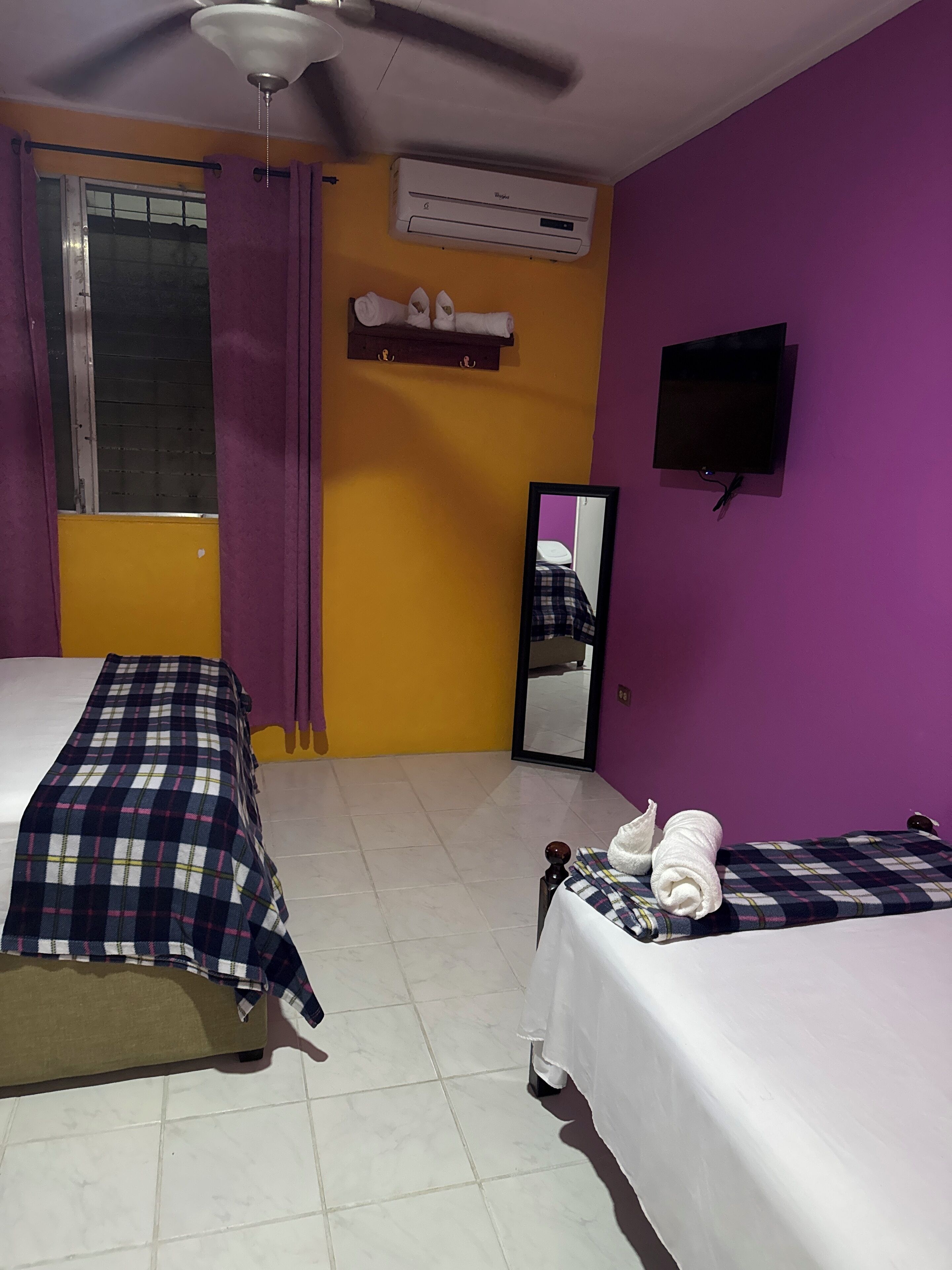 Une chambre avec deux lits, un ventilateur de plafond, un appareil de climatisation, une télévision et un miroir au mur.