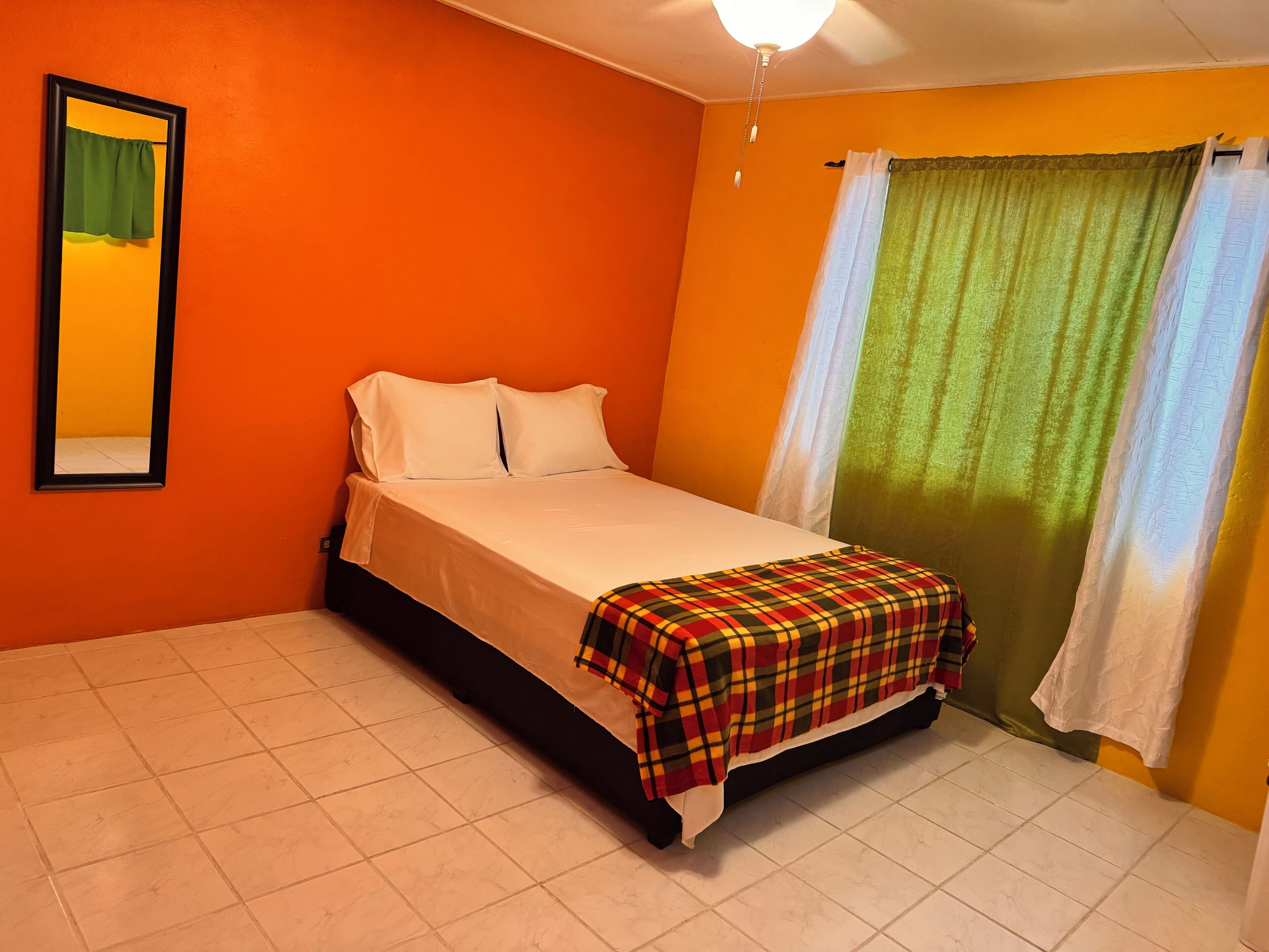A room with an orange wall, a bed with white bedding, a plaid blanket, a window with green curtains, and a ceiling light.