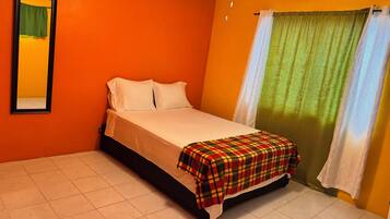 A room with an orange wall, a bed with white bedding, a plaid blanket, a window with green curtains, and a ceiling light.