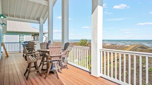 Une terrasse en bois avec des meubles d’extérieur, une balustrade blanche et une vue sur la plage et l’océan.