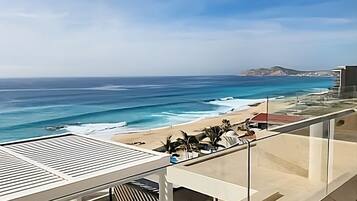 Balcón con vista al océano y a la playa, con mobiliario exterior y baranda de vidrio.