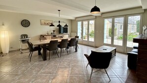 A spacious dining area with a large table and chairs, a kitchen in the background, and large windows offering a view of the outdoors.
