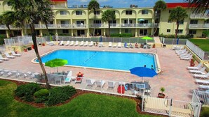 A hotel with a swimming pool surrounded by lounge chairs and umbrellas, a red-roofed building, and a fenced area with greenery. - Oceanfront End-Unit | Pool View, Washer/Dryer (St. Augustine Beach)