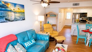 A living room with a blue sofa, a small table, a chair, a ceiling fan, and a kitchen in the background. - Oceanfront End-Unit | Pool View, Washer/Dryer (St. Augustine Beach)