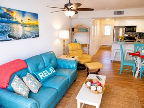 A living room with a blue sofa, a small table, a chair, a ceiling fan, and a kitchen in the background. - Oceanfront End-Unit | Pool View, Washer/Dryer (St. Augustine Beach)