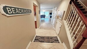 A hallway with a 'BEACH HOUSE' sign, a wooden staircase, and a patterned rug. - Ocean-View Condo | Pool + Balcony | Crescent Beach (St. Augustine Beach)