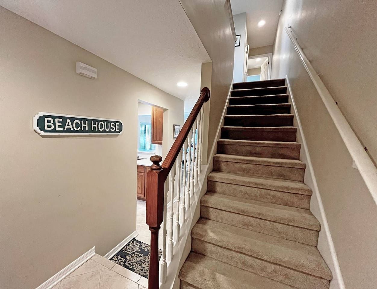 A staircase with a wooden handrail and beige carpeted steps leading to a landing. A sign on the wall reads "BEACH HOUSE."