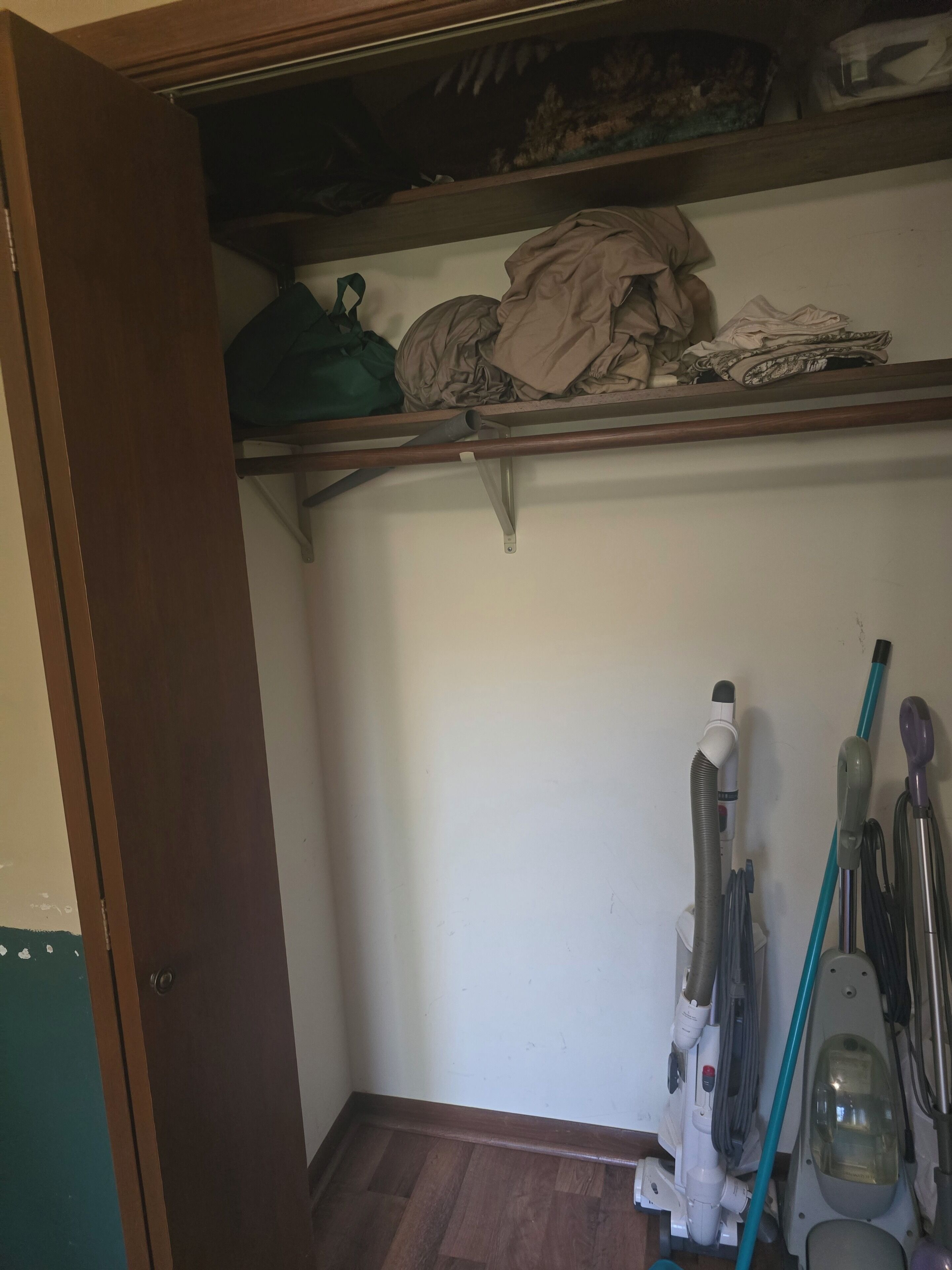 Una habitació amb una porta de fusta, una estant amb roba doblegada i un aspirador al sòl.