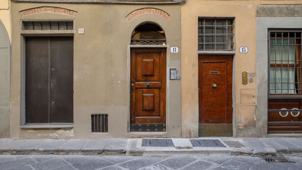 Tres puertas de madera en fila, con placas numeradas, frente a una pared color beige y una calle empedrada.