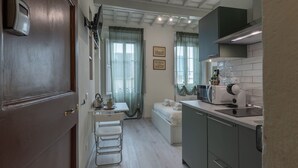 Una cocina compacta con microondas, una mesita y una silla. Hay una cama con colchón blanco y una ventana con cortinas translúcidas.