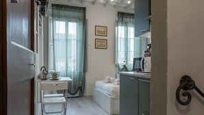 Un dormitorio compacto con cama, un escritorio pequeño y una silla. Se ve una ventana con cortinas, un estante en la pared con objetos y una puerta que da a otra habitación.