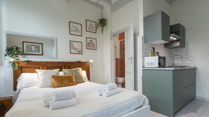 Un dormitorio con cama, almohadas y mesita de noche. Se ve un baño con inodoro y ducha a través de una puerta abierta. La habitación tiene una kitchenette con microondas y armarios.