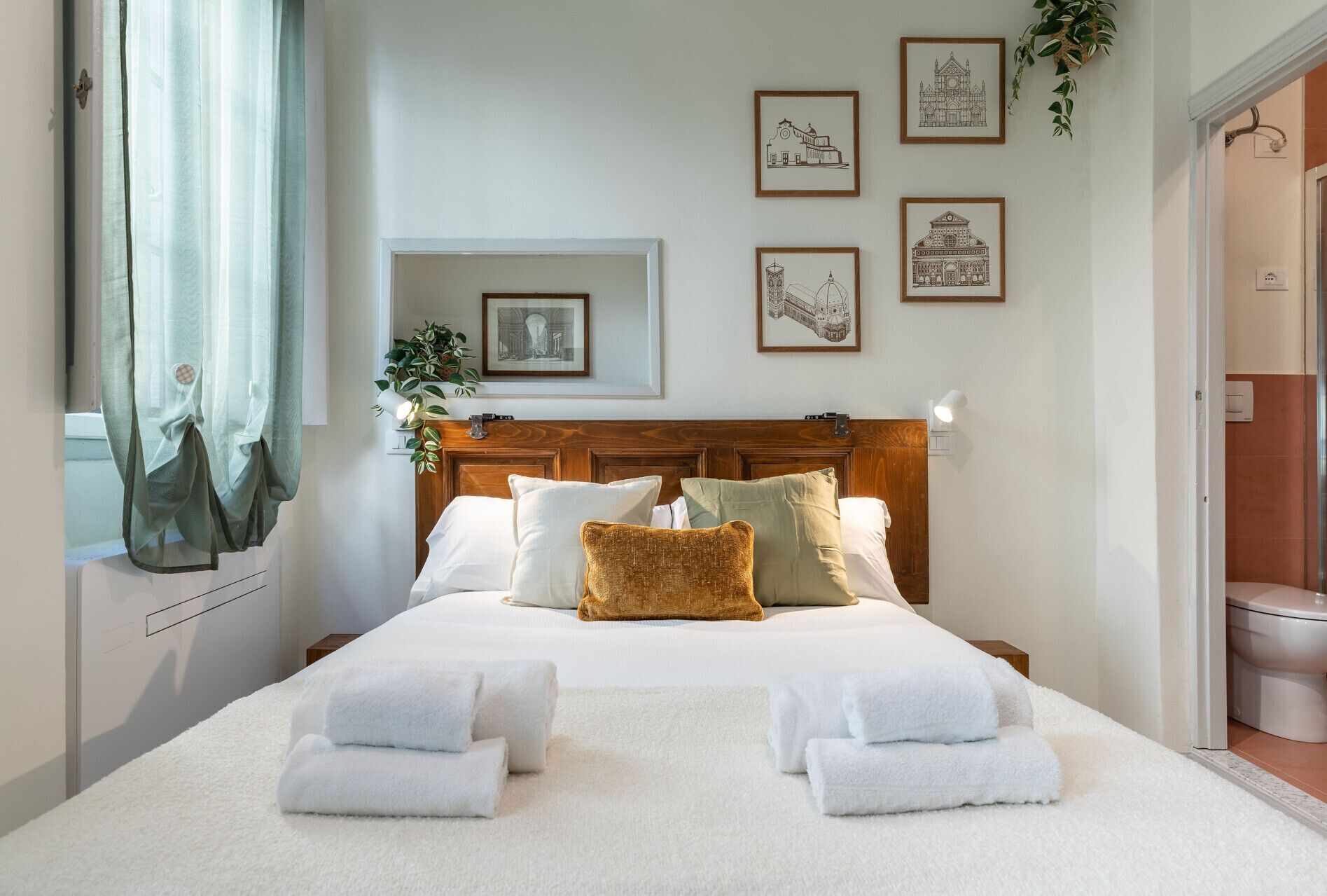 A neatly made bed with white linens and towels, a wooden headboard, and a wall with framed architectural drawings.
