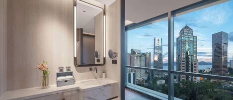 Un baño moderno con un espejo grande, lavamanos dobles y vistas a un paisaje urbano a través de amplias ventanas.