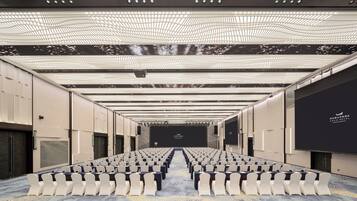 Un salón de conferencias con filas de sillas, un escenario y una pantalla grande.