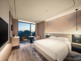 A modern hotel room with a large bed, a desk, a chair, and a view of the cityscape.