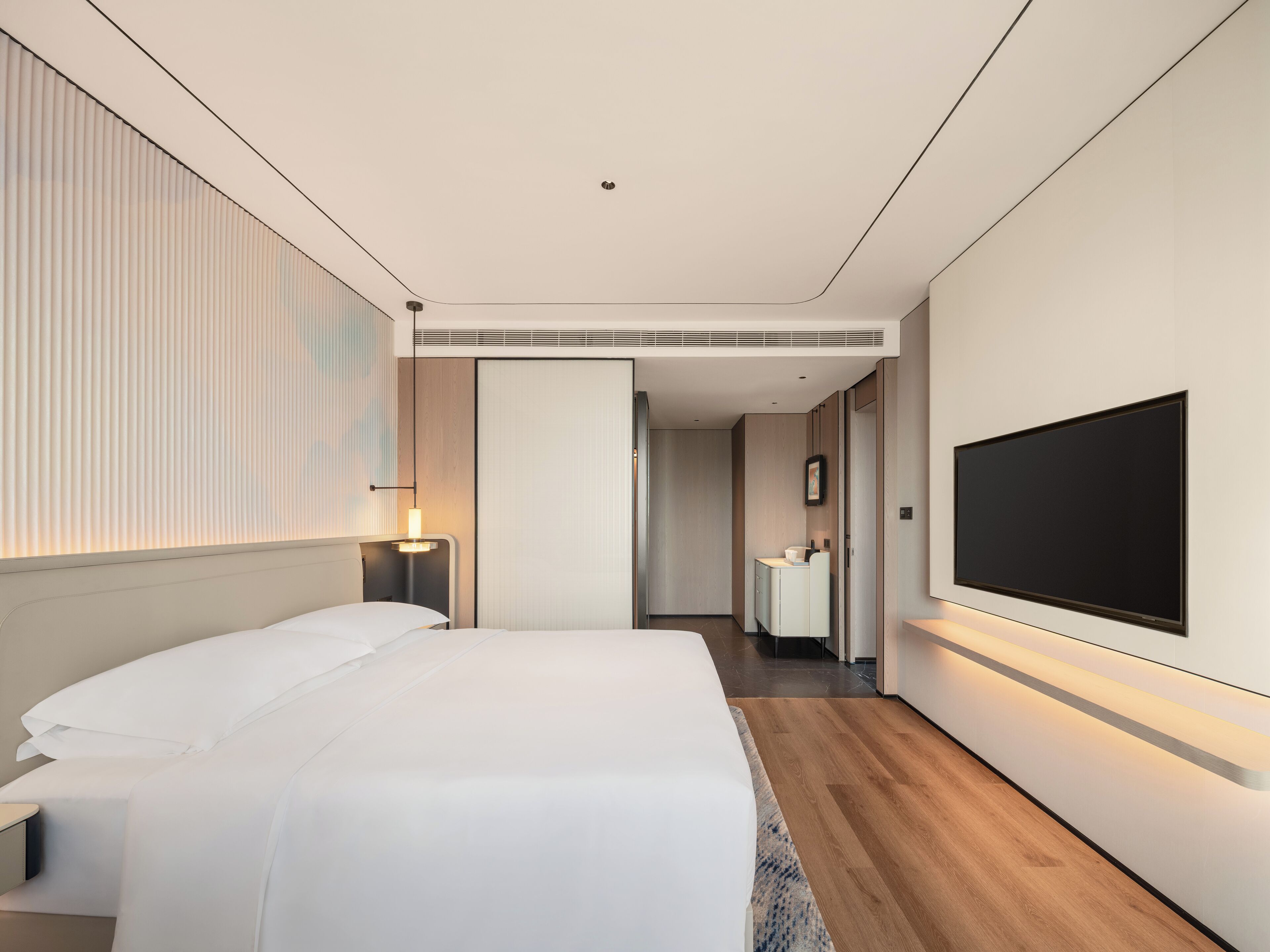 A modern hotel room with a large bed, a flat-screen TV mounted on the wall, a bedside table, and a nightstand.