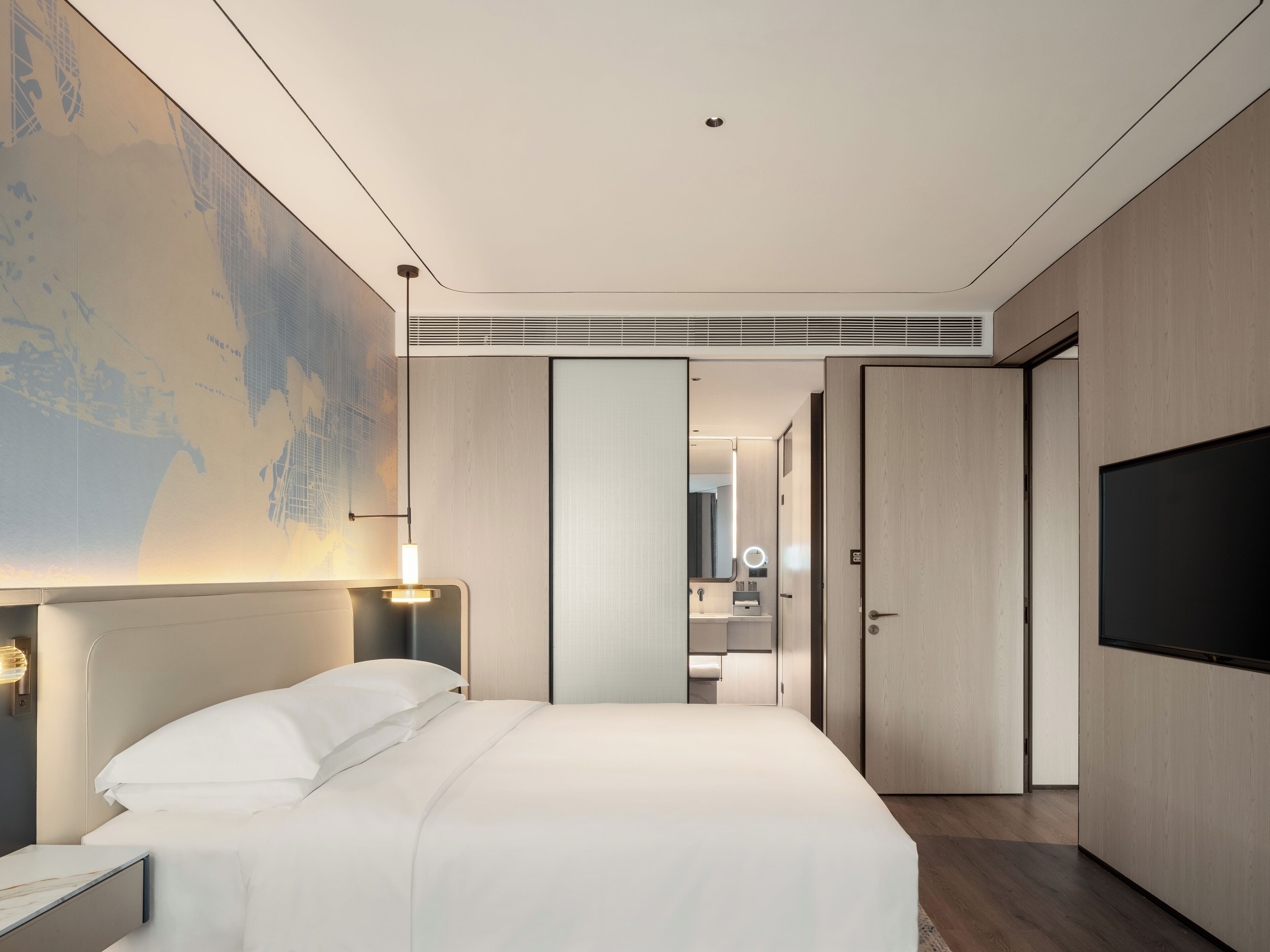 Une chambre d’hôtel moderne dotée d’un grand lit, d’un téléviseur à écran plat, d’une table de chevet, d’une lampe fixée au mur et d’un grand tableau abstrait.